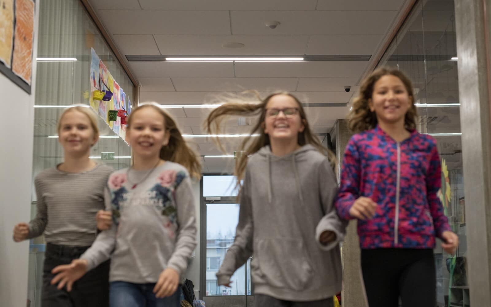 Neljä lasta juoksee iloisina rinnakkain koulun käytävällä.